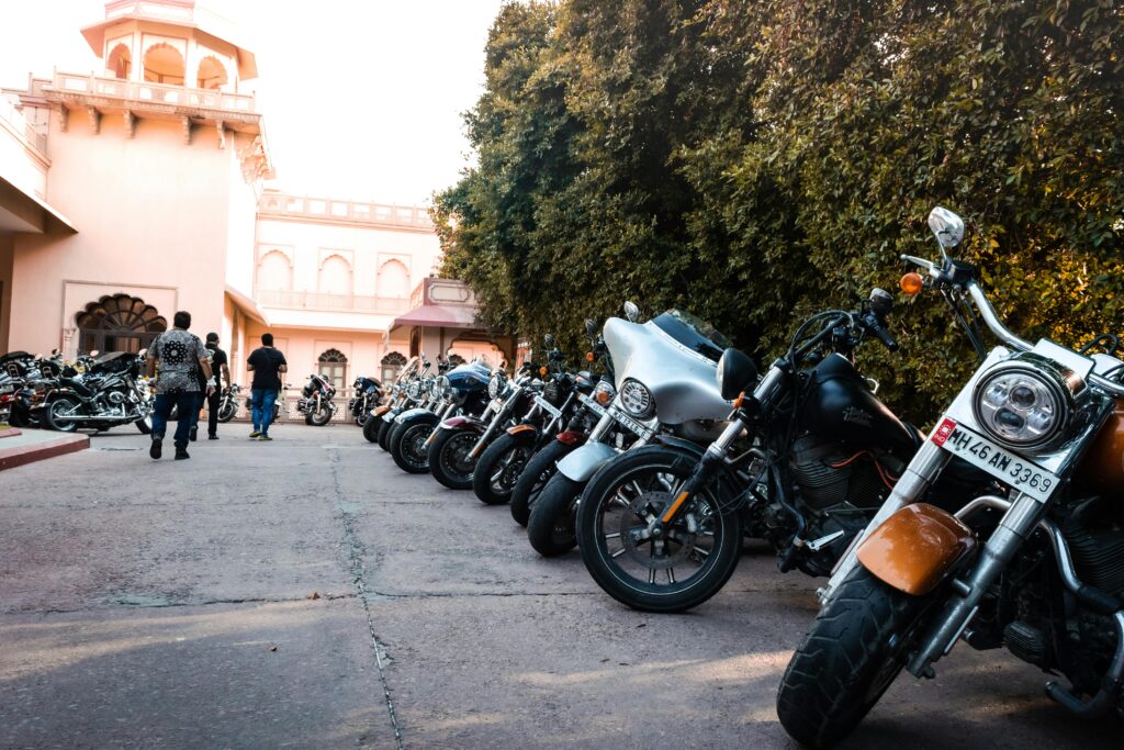 A row of motorcycles parked outside a historic building with trees and people walking.