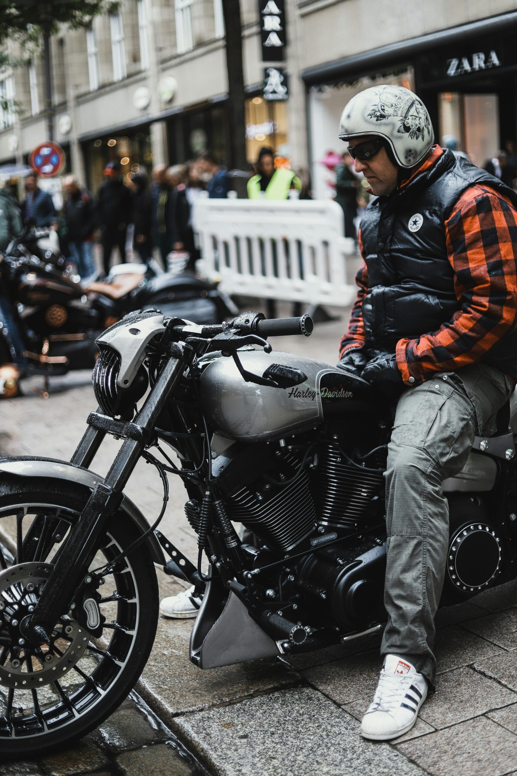 Biker in puffer vest and plaid shirt seated on Harley Davidson motorcycle on bustling Hamburg street.